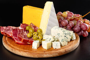 Wooden kitchen board with sliced delicatessen cheeses, ham, olives and grapes - image #475929 gratis